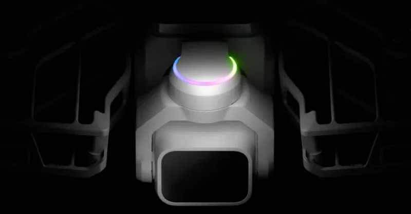 A close-up, centered view of a drone camera gimbal with a glowing, multi-colored LED ring, flanked by two dark, structural components.