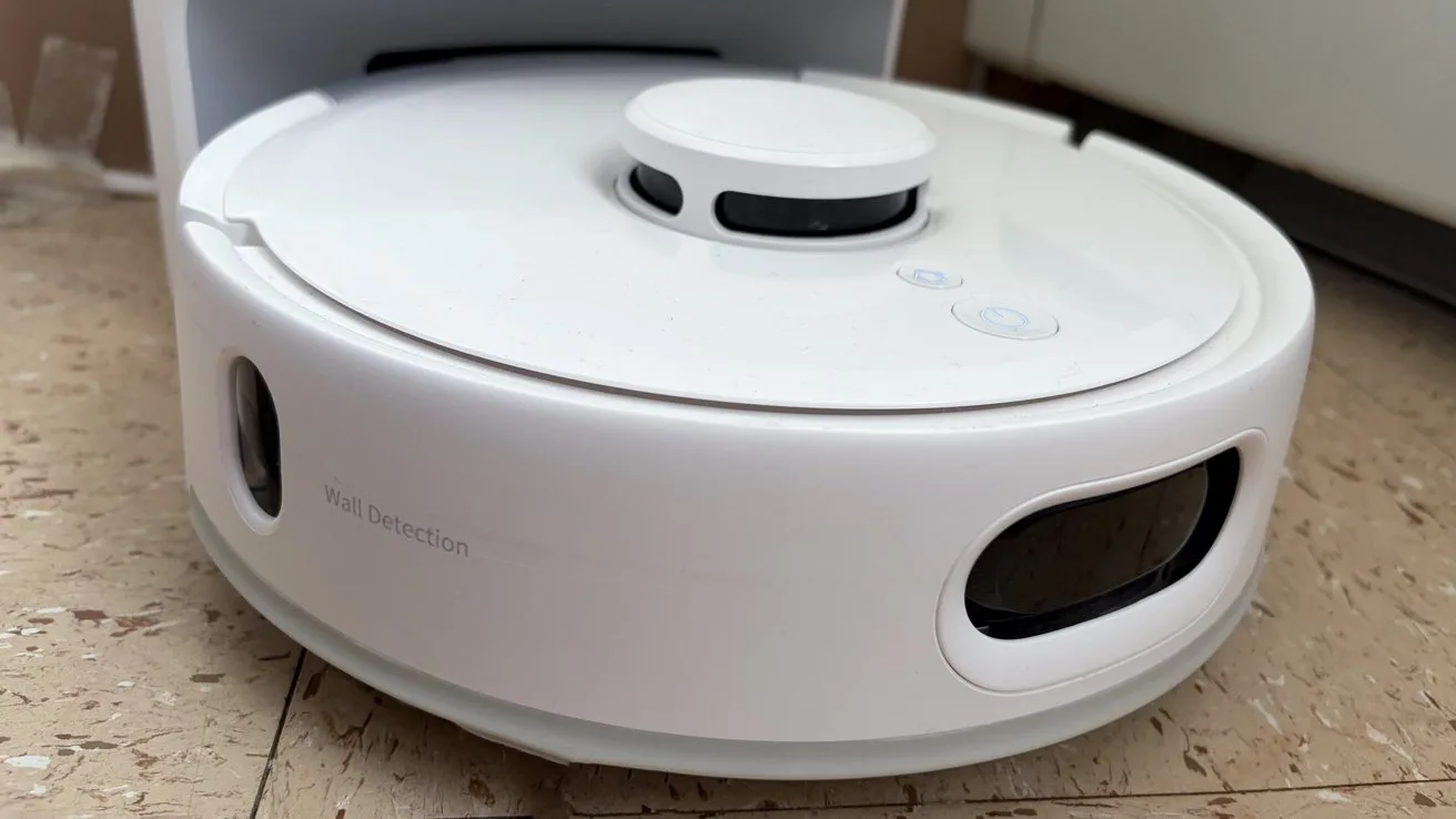 White circular robot vacuum cleaner docked on a hard floor, showing its front sensor window, side vents, and raised lidar-style sensor on top