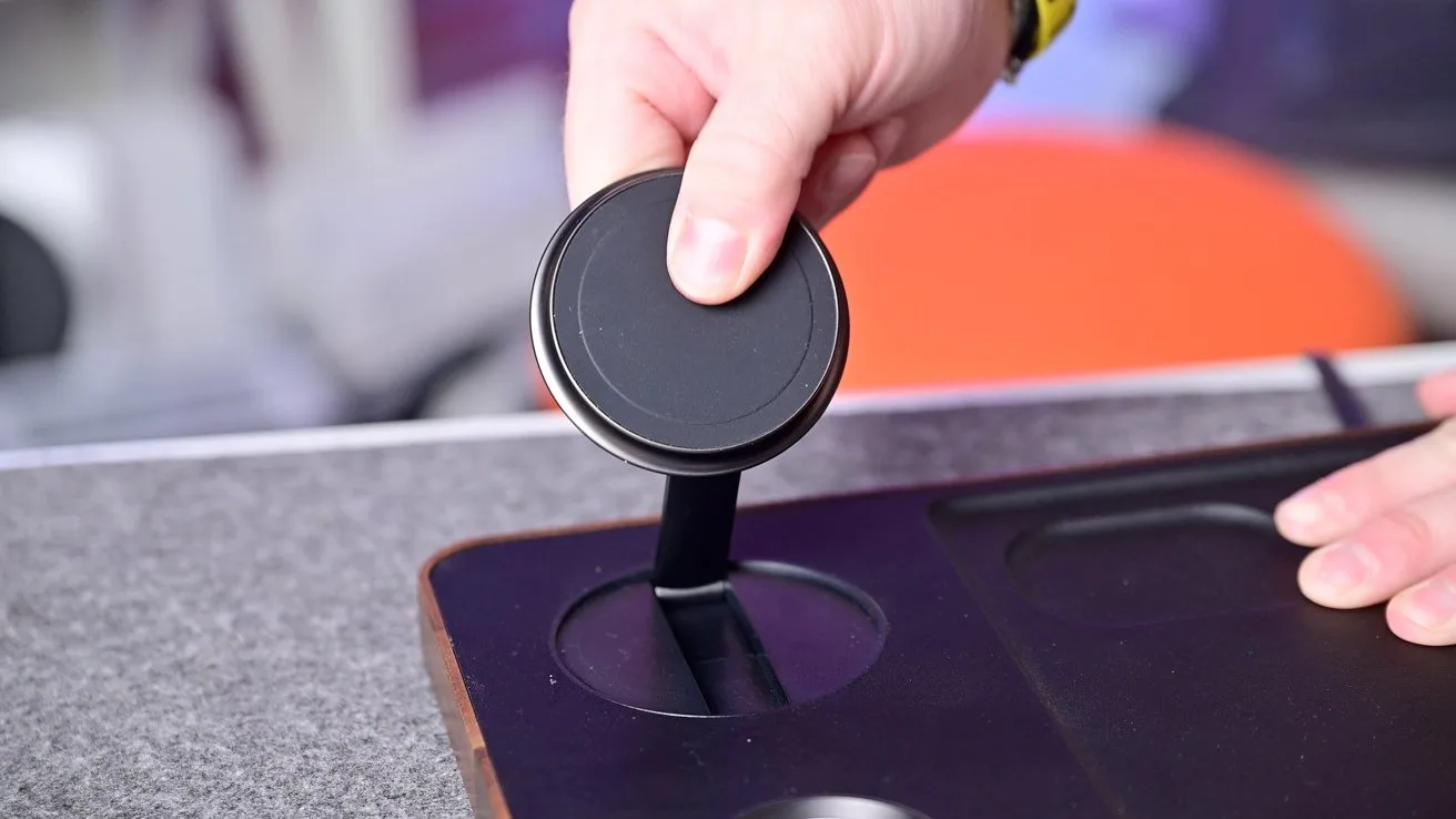 Hand lifting a round black wireless charger from a recessed slot in a desk organizer, with a gray desktop surface and blurred colorful background
