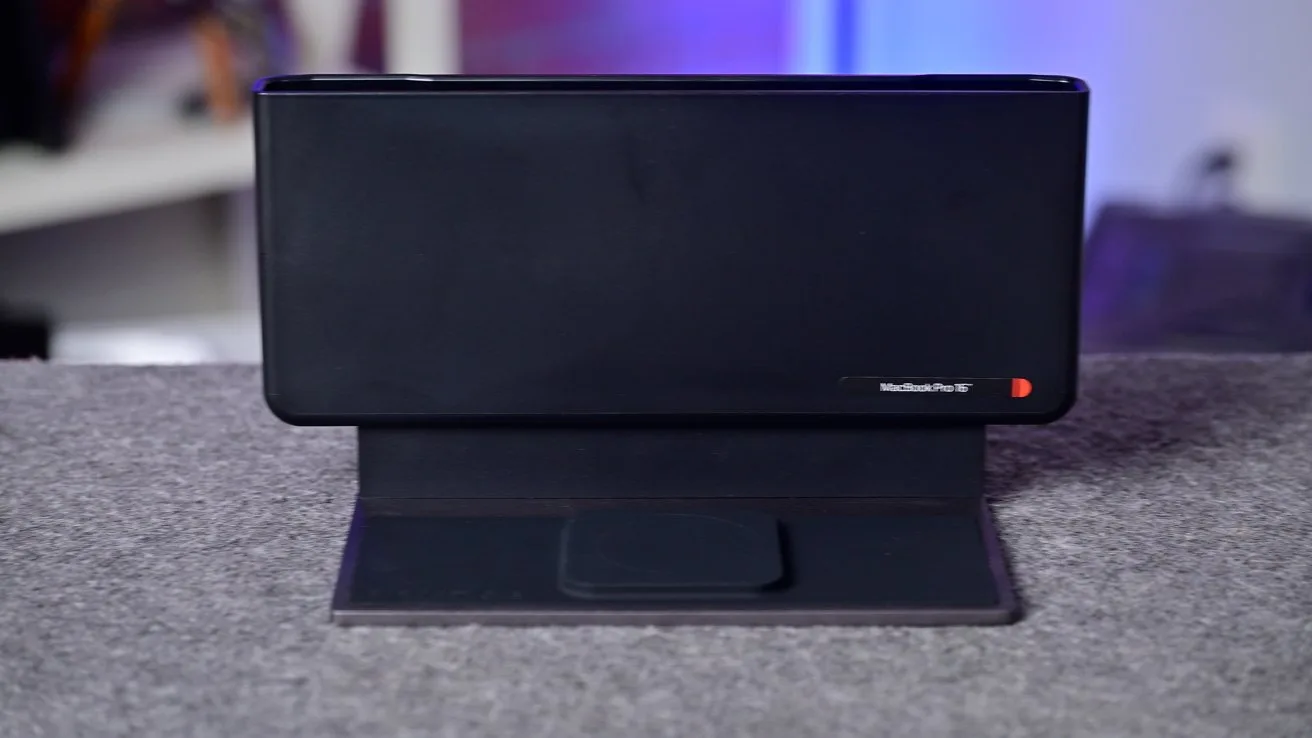 Black rectangular electronic device standing vertically on a folded cover or stand atop a gray surface, with a small red accent and blurred shelves and lights in the background