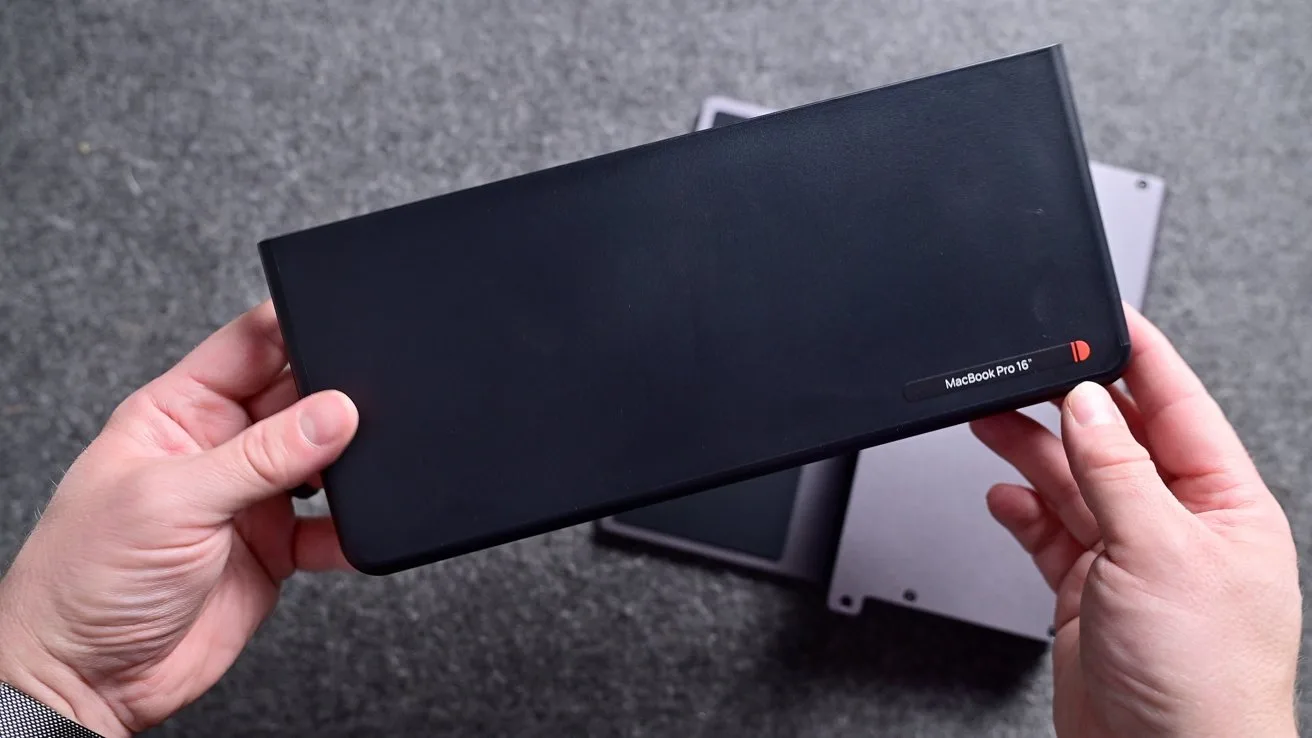Hands holding a long black handheld device with rounded corners above two rectangular electronic gadgets on a gray surface