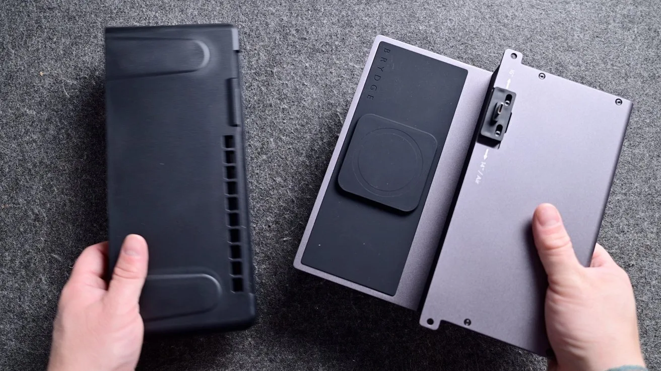 Two hands holding two rectangular electronic devices on a gray surface, one black with vent-like grooves, the other silver with a large black pad and mounting bracket
