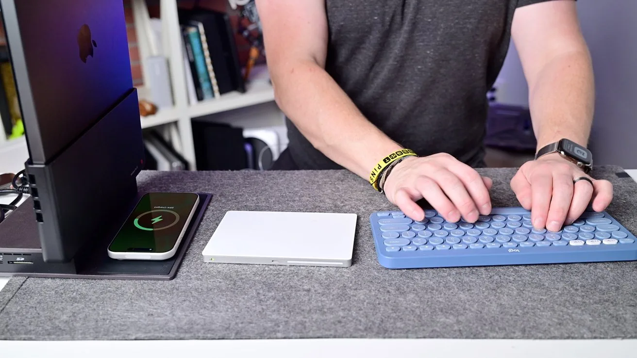 Person typing on a blue wireless keyboard at a tidy desk with a smartphone on a charging pad, a trackpad, a vertical laptop stand, and shelves of books in the background