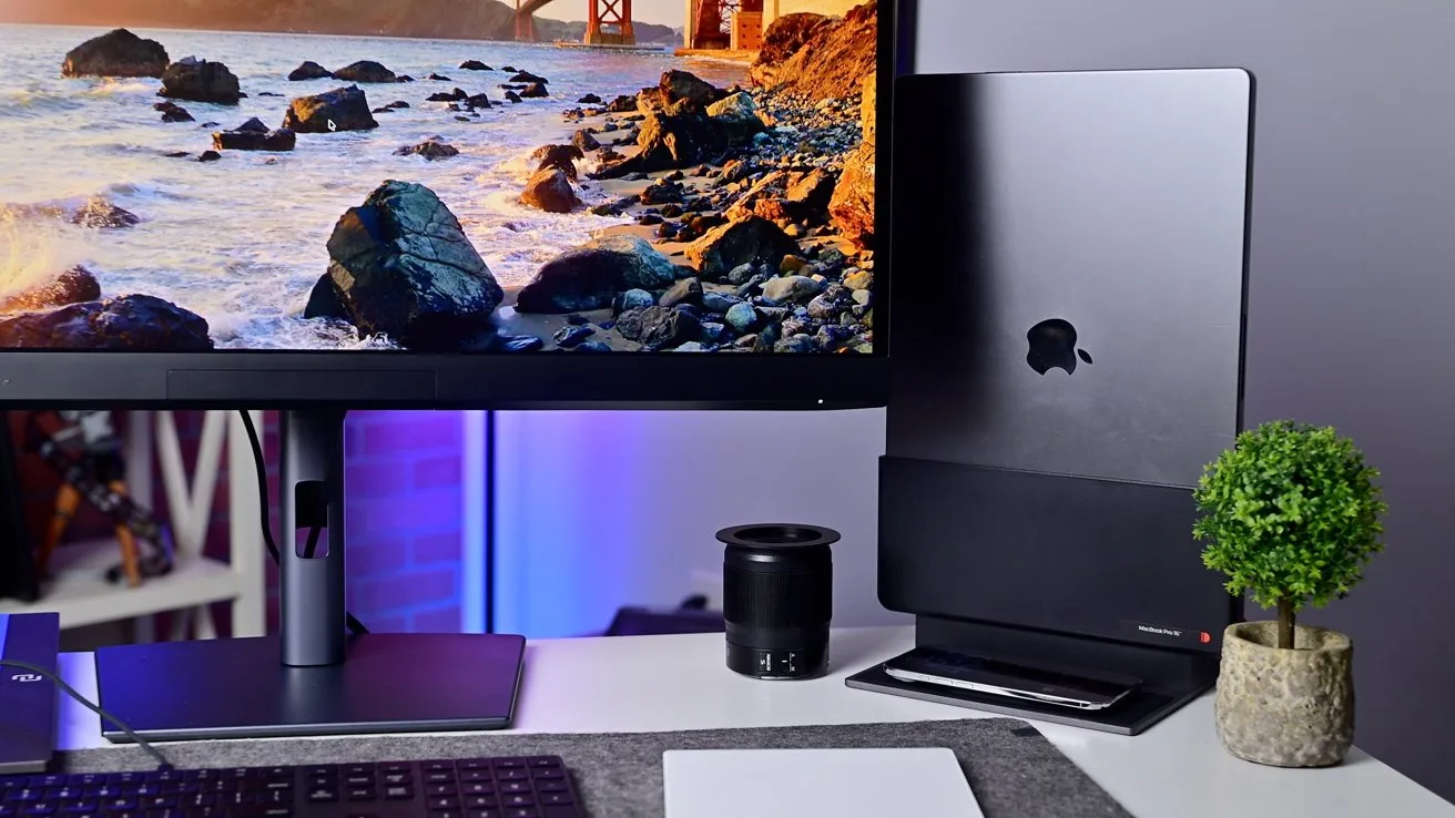 Modern desk setup with ultrawide monitor showing a rocky coastline, vertical closed MacBook on a stand, small potted plant, keyboard, mousepad, and a black speaker on a clean workspace
