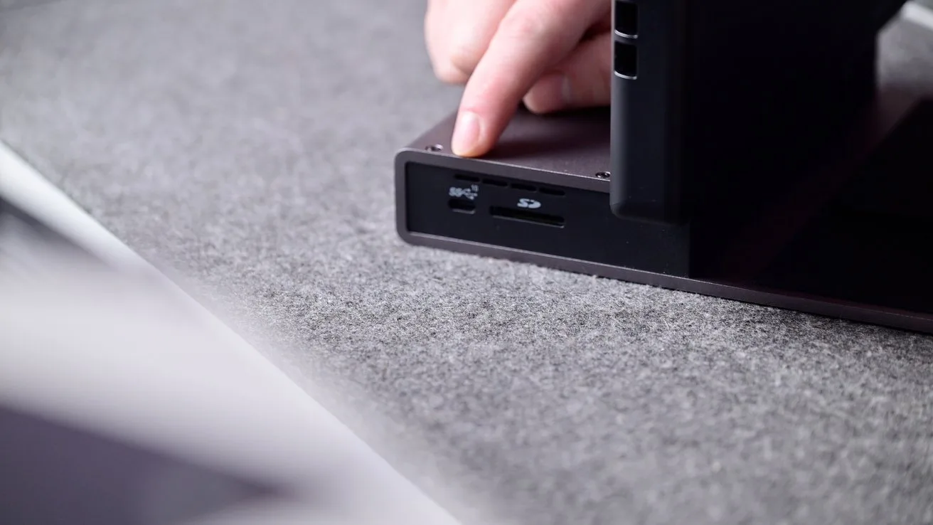 Hand pointing to USB and memory card slots on the side of a dark electronic device resting on a gray fabric surface