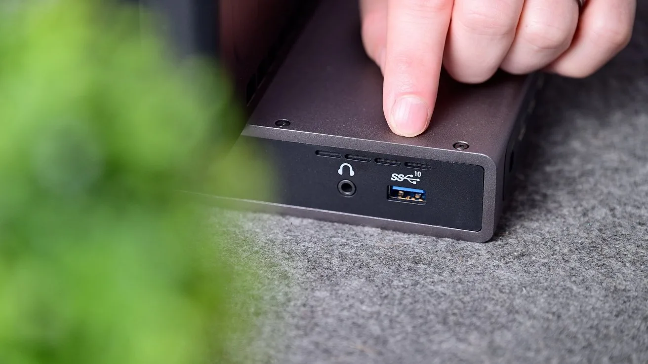 Hand pressing a button on a small black electronic device with headphone jack and blue USB 3 port on the front, resting on gray fabric near a blurry green plant