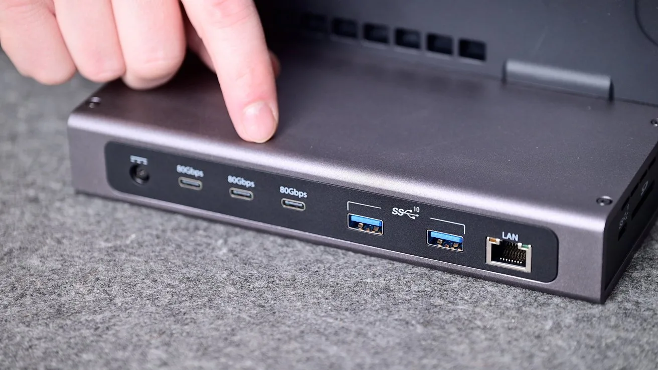 Person pointing at the back of a compact computer docking station showing multiple USB-C ports, two blue USB-A ports, and a single Ethernet LAN port on a gray surface