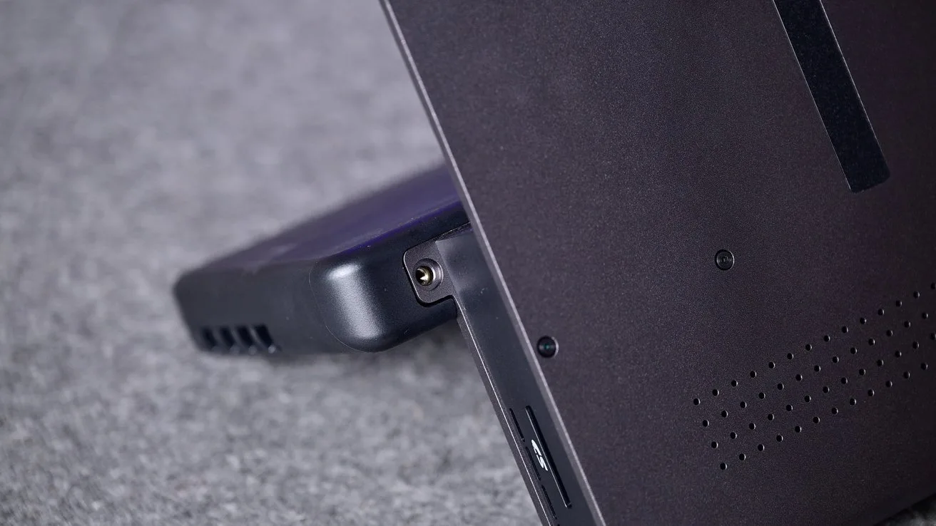 Close-up of a tablet or 2-in-1 laptop propped on its kickstand, showing side ports, ventilation holes, and a connected accessory, resting on a soft gray surface