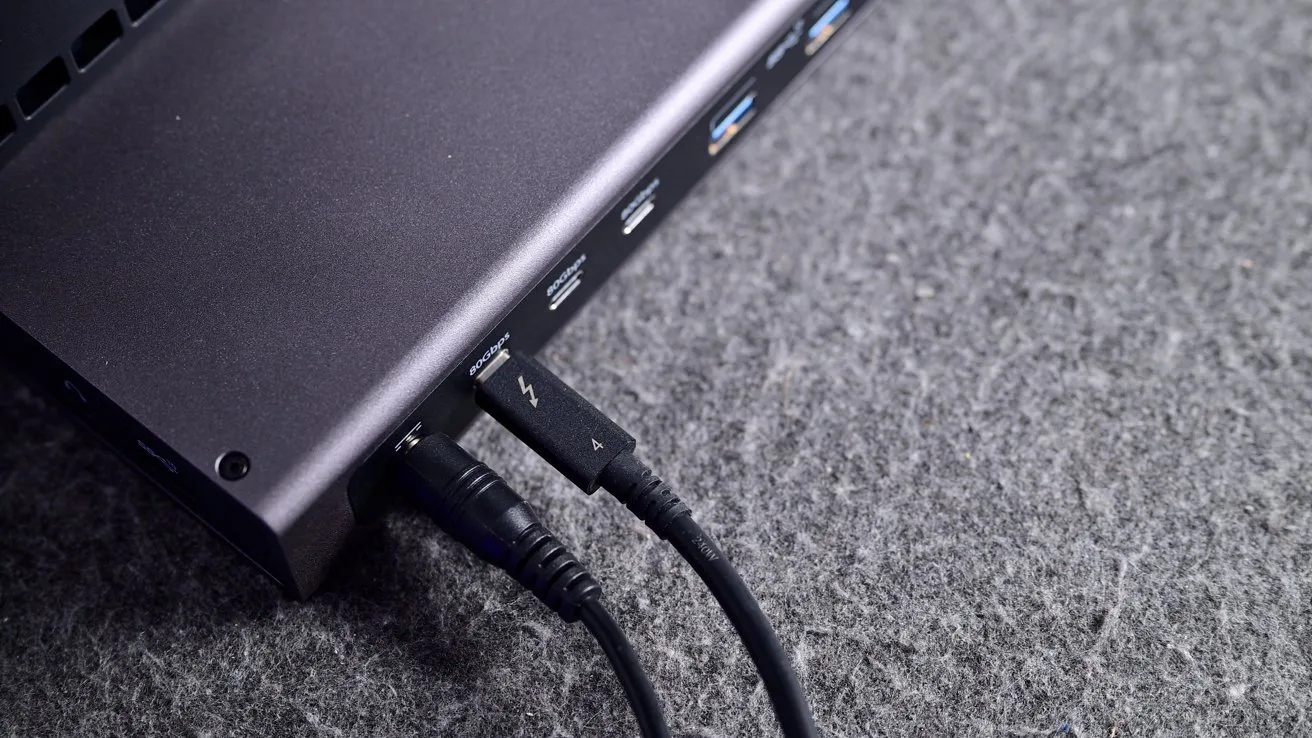 Close-up of a laptop's side ports on gray fabric, showing multiple USB ports with cables plugged in, including a Thunderbolt USB-C cable and an audio or power cable