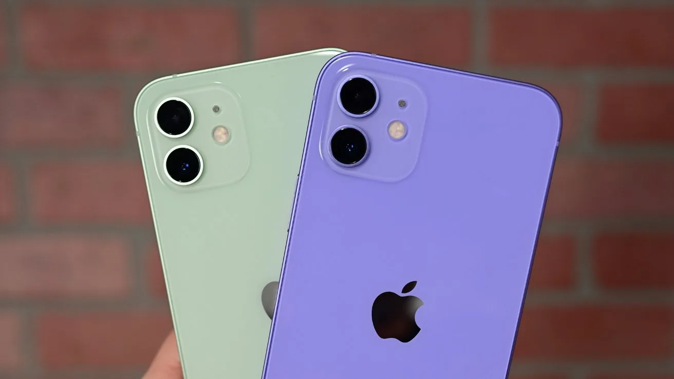 Two iPhones held up, one light green and one purple, both showing their backs with dual cameras and Apple logos, against a blurred red brick wall background