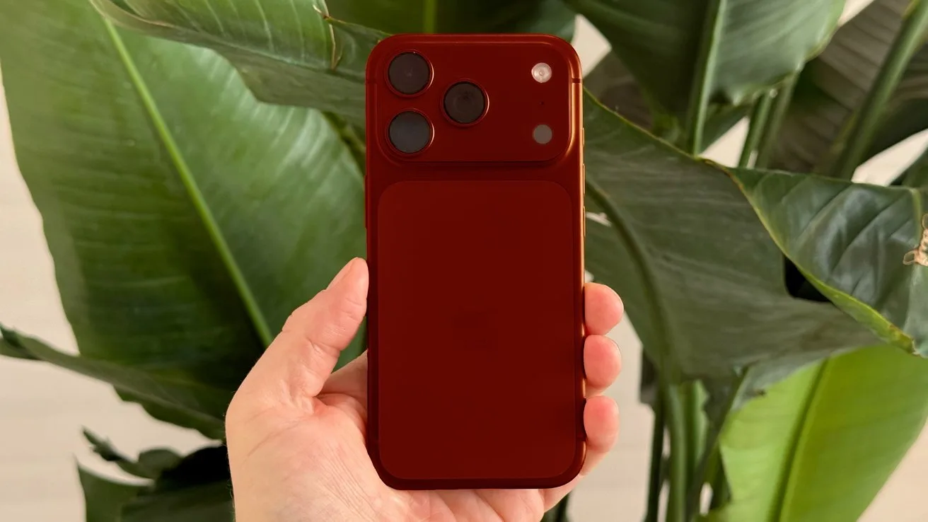 Hand holding a red smartphone with three rear camera lenses, positioned vertically in front of large green tropical leaves in soft natural light