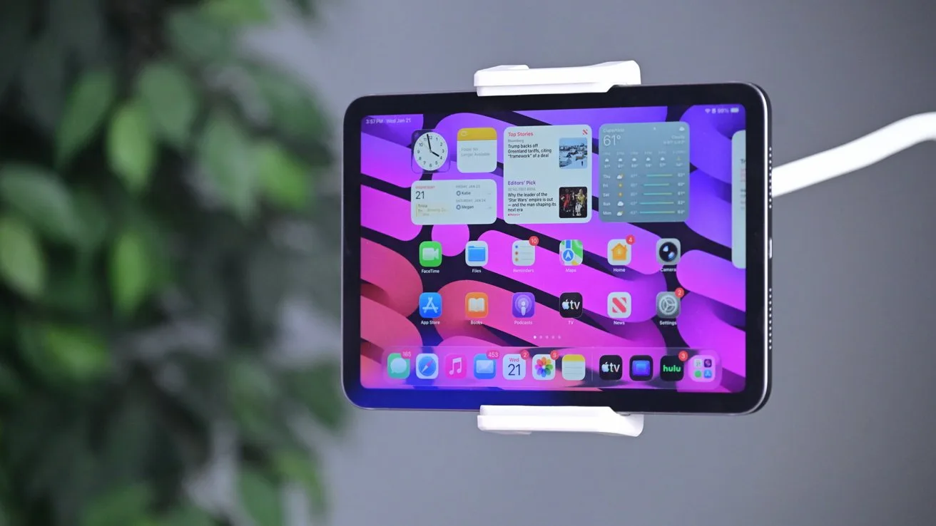 Tablet in a white clamp mount displaying a colorful pink and purple wavy home screen with app icons and widgets, set against a blurred indoor background with green plant leaves