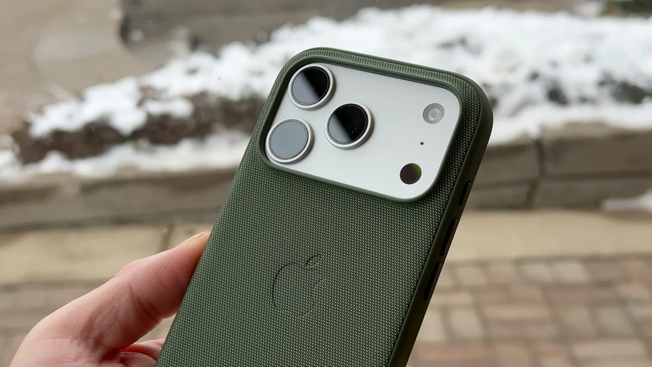 Hand holding an iPhone with a green textured Apple case, showing dual rear cameras and flash, outdoors near a snow-covered sidewalk and stone edging