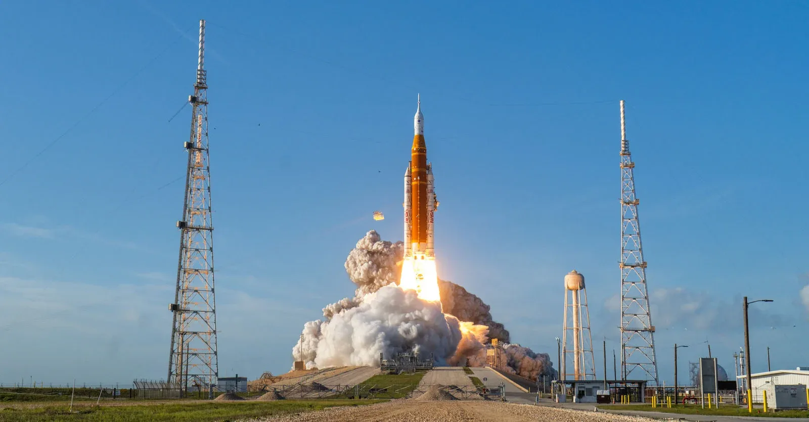 A large orange and white rocket launches into a clear blue sky, surrounded by a massive cloud of smoke and two tall towers.