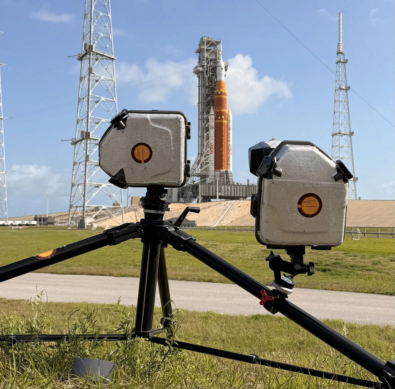Two cameras in protective, insulated housings are mounted on a tripod in a grassy field, with a large rocket on a launch pad in the background.