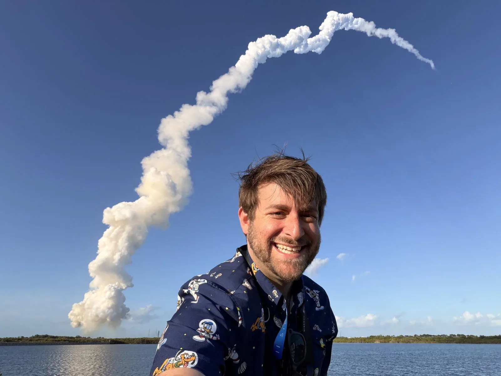 A smiling man wearing a patterned shirt poses for a selfie in front of a rocket launch plume rising into a clear blue sky.