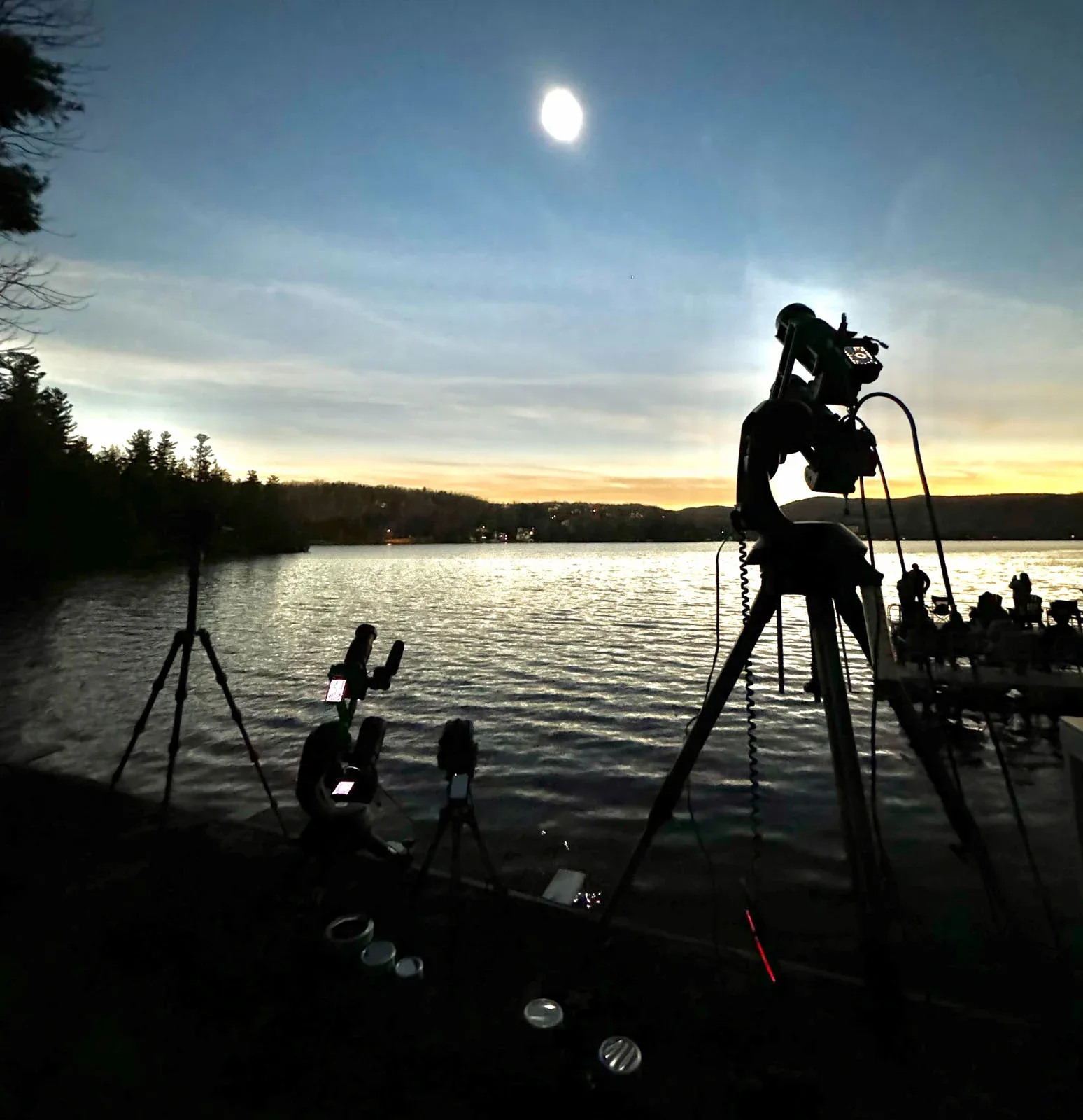 Several tripods with mounted cameras and telescopes are set up along a lakeshore at dusk under a bright moon.