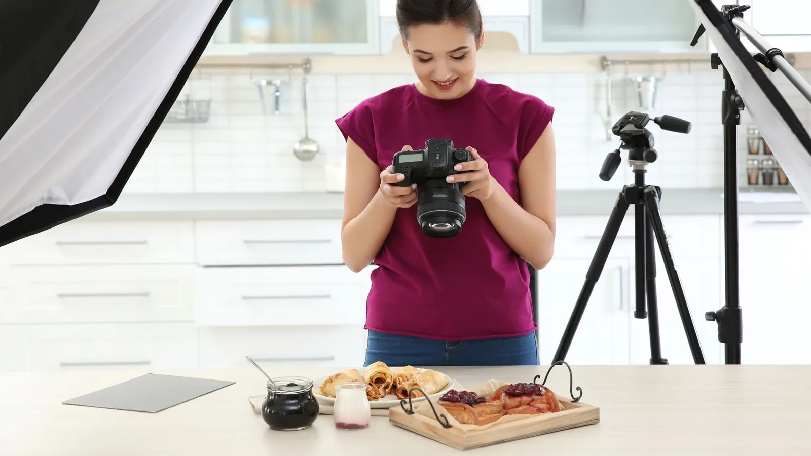 A photographer in a kitchen setting, adjusting the camera while photographing a plate of food on a table, with soft lighting equipment in the background.