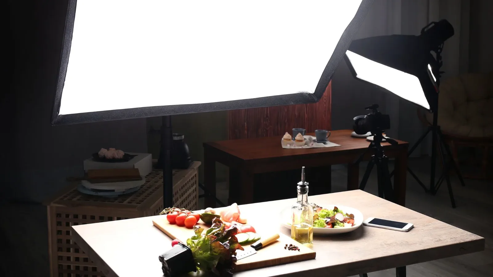 A food photography studio table set up with ingredients for a fresh salad, surrounded by professional lighting equipment, and a smartphone nearby.
