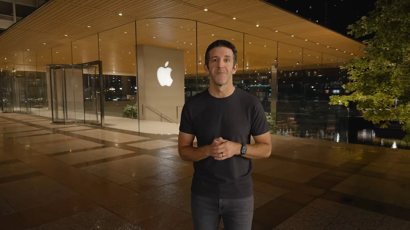 Man in dark tshirt stands outside a modern glass Apple Store at night, hands clasped, with large glowing Apple logo behind him and wet reflective pavement in foreground