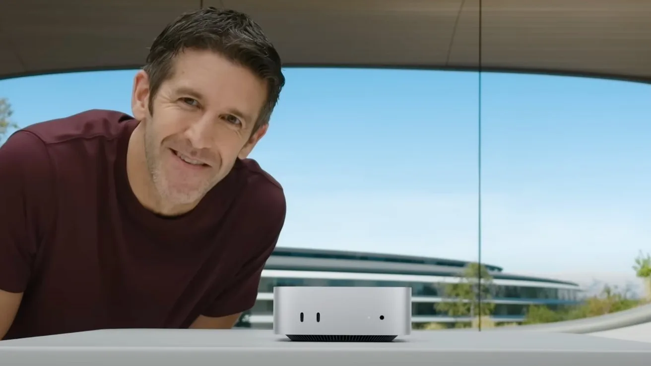 Smiling man in a maroon shirt leans over a table displaying a small silver desktop computer, with a modern glass building and blue sky in the background