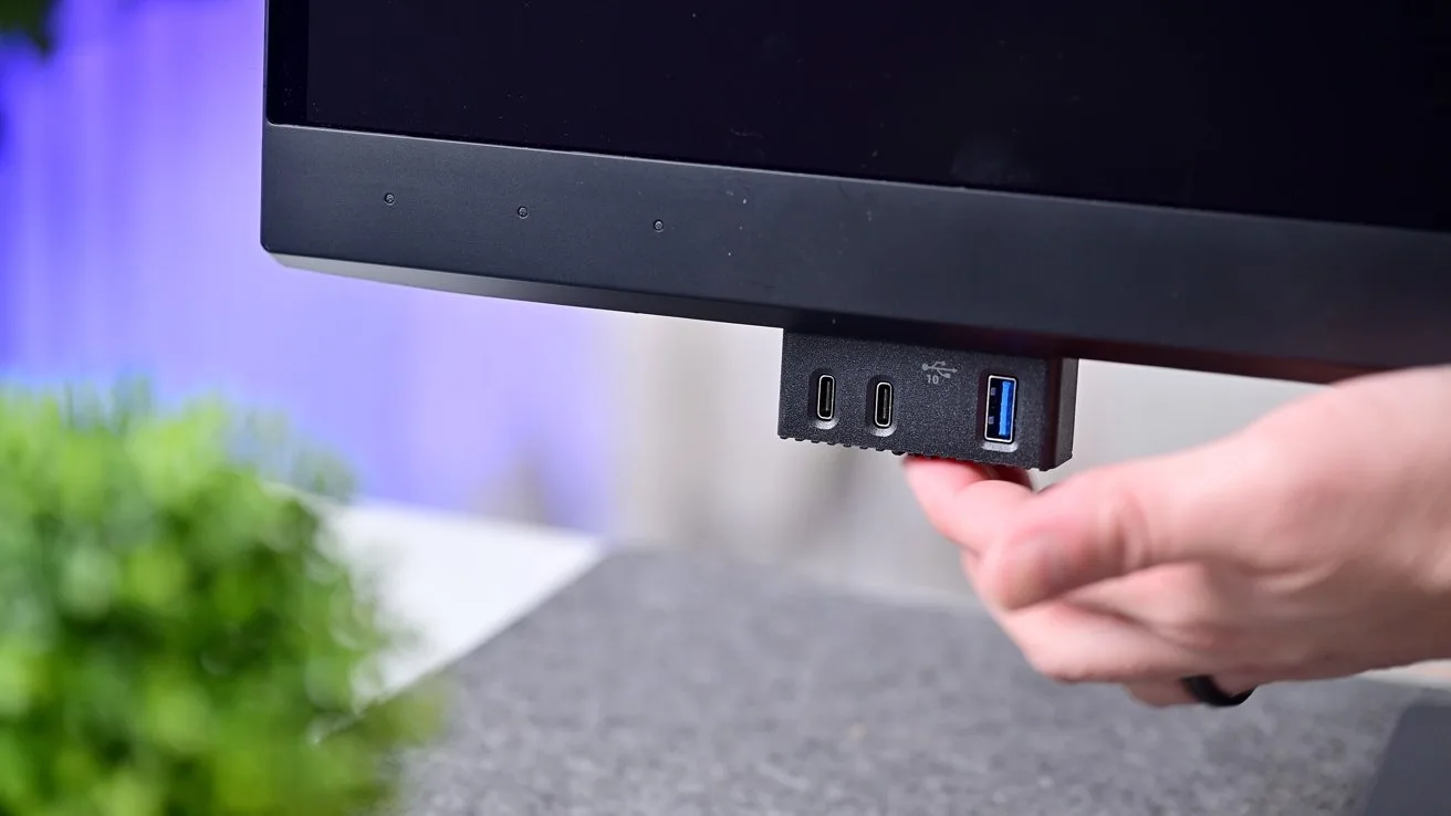 Hand pulling a small USB hub from the bottom edge of a monitor, showing USB-C and USB-A ports, with a blurred green plant and desk surface in the background