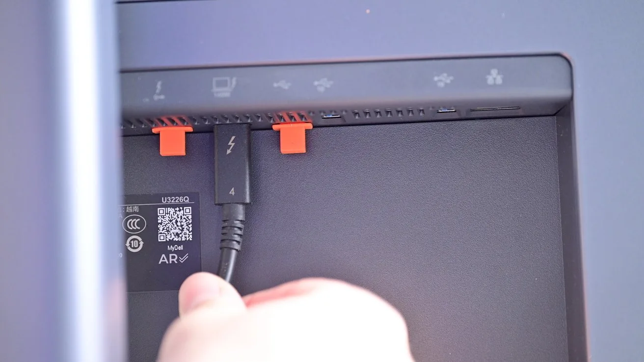 Hand plugging a Thunderbolt cable into a port on the back of a monitor or computer, near orange clips and a QR code label with small icons above the ports