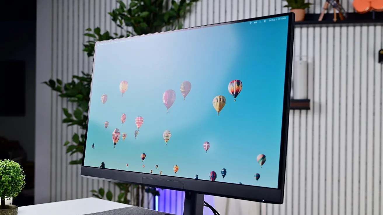 Modern computer monitor on a desk displaying colorful hot air balloons in a blue sky, with indoor plants and vertical white wall panels in the softly lit background