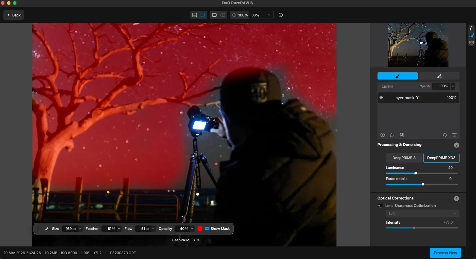 A person in dark clothing takes a photo of a vivid red night sky filled with stars, while editing the image on a computer screen using photo editing software. A leafless tree is visible on the left.