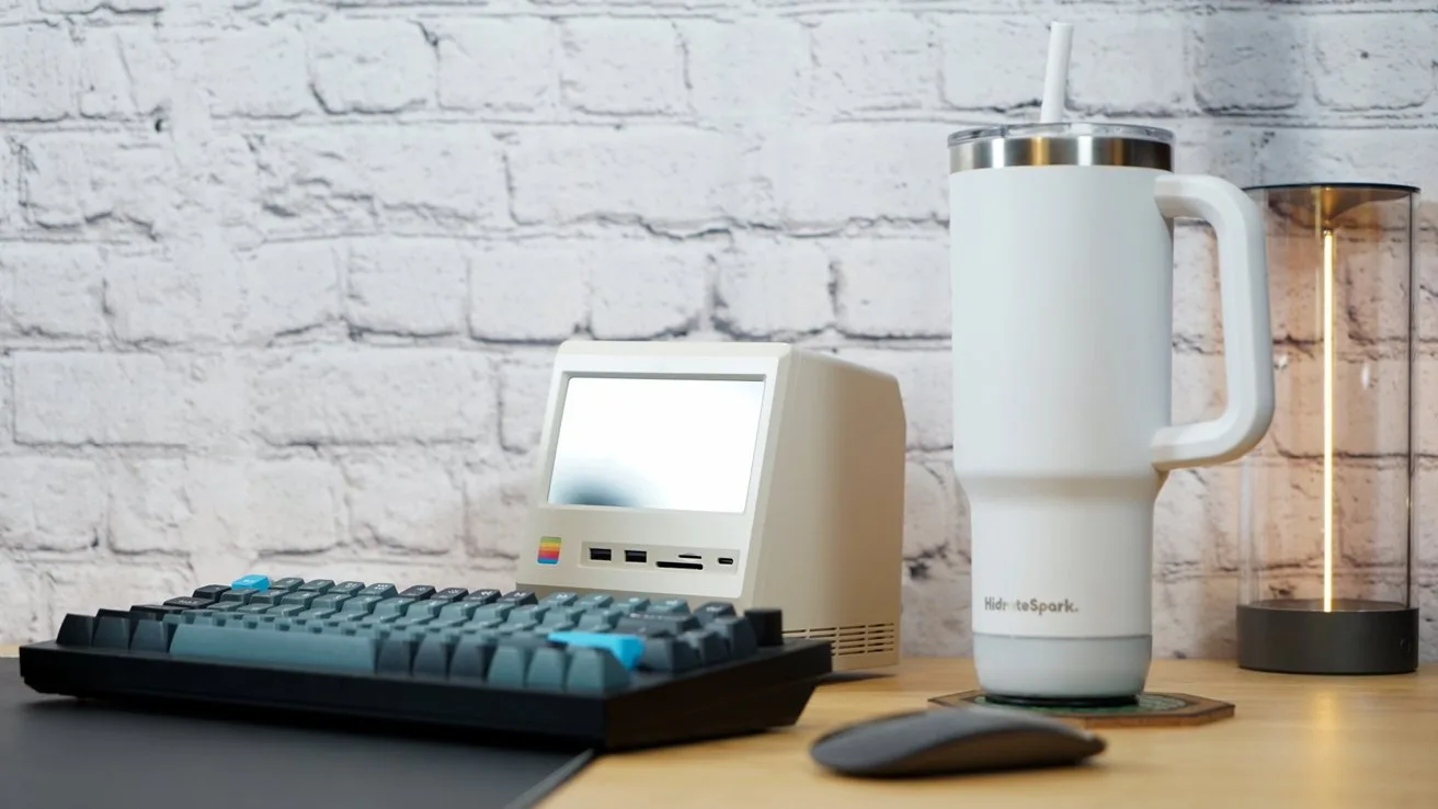 Retro-style small computer, gray keyboard, tall white tumbler with straw, glowing LED lamp on wooden table, white brick wall background.
