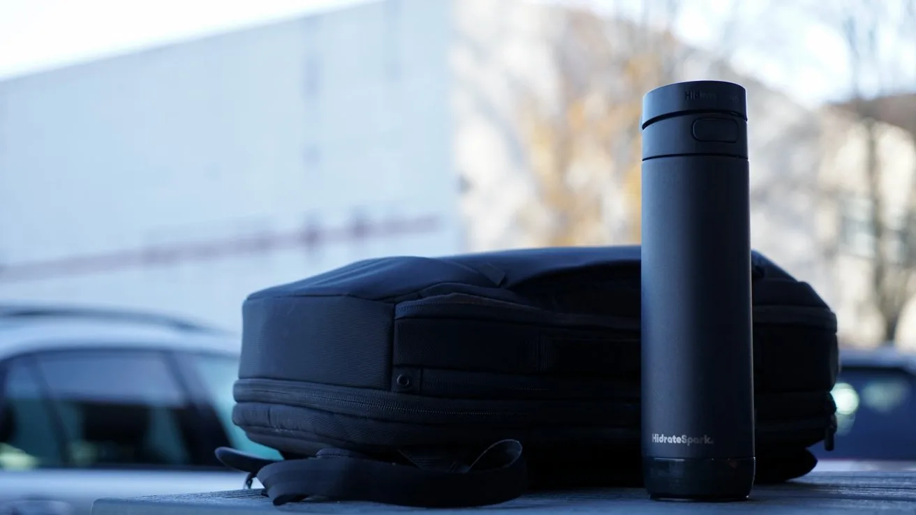 A black tumbler bottle and a dark backpack rest on a concrete surface with blurred outdoor background.
