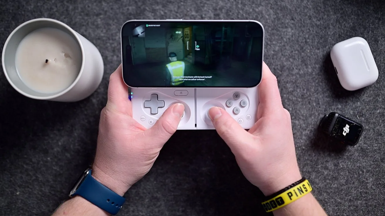 Hands holding a handheld gaming device displaying a game. Nearby are a candle, earbuds case, and a small device.