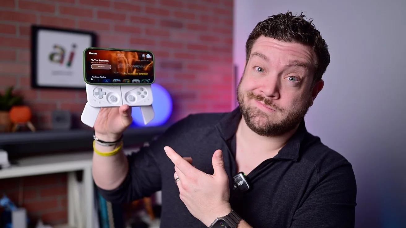 Man in dark shirt holding a smartphone with detachable game controller, smiling and pointing at it in a room with brick wall background.
