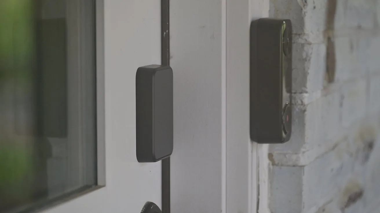 Close-up of a closed glass door with a rectangular smart sensor or contact sensor mounted on the door frame and a matching piece on the adjacent brick wall