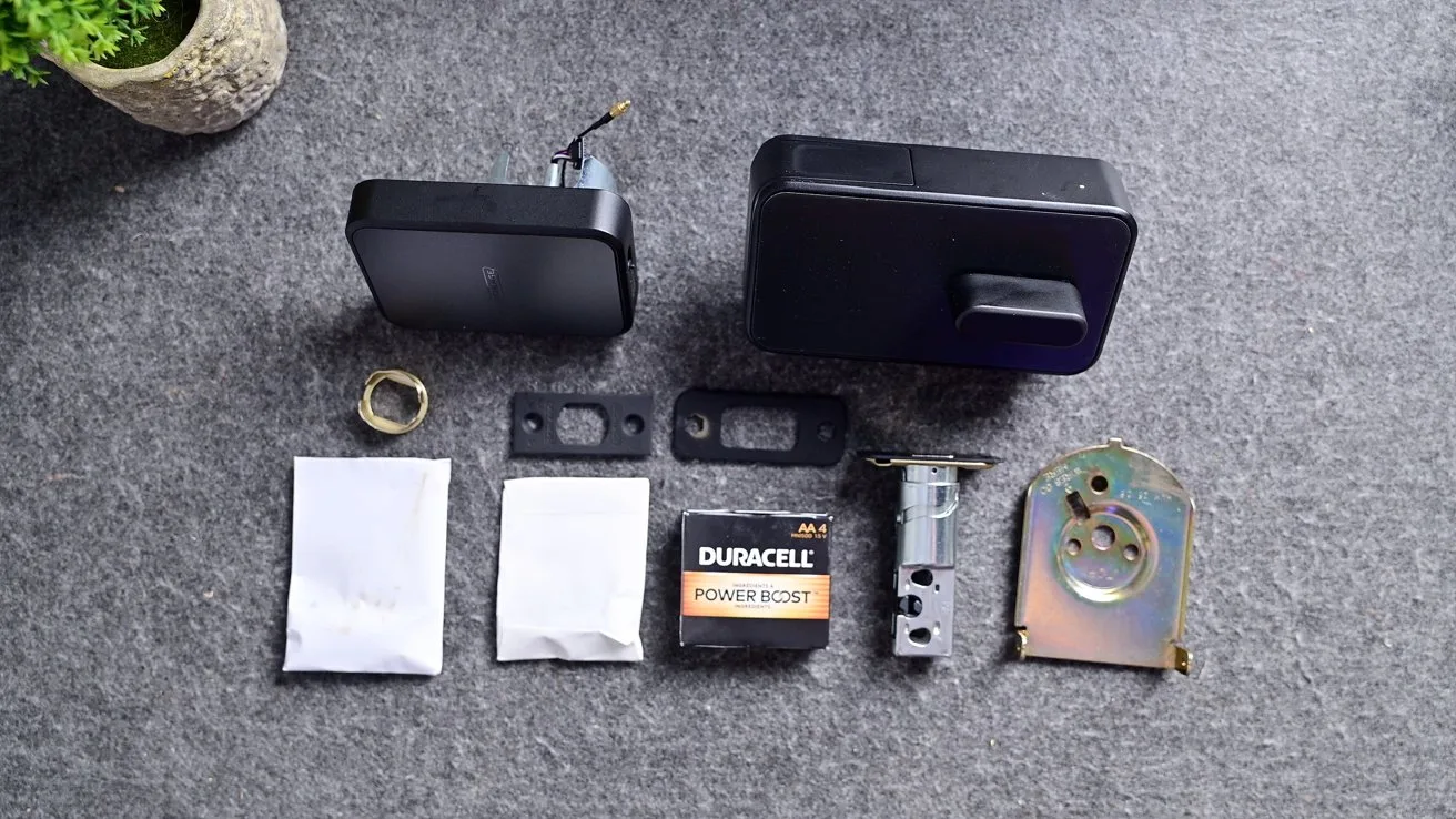 Electronic door lock components laid out on gray surface, including black lock units, metal latch and strike plates, screws in packets, mounting hardware, and a Duracell battery, with plant partially visible.