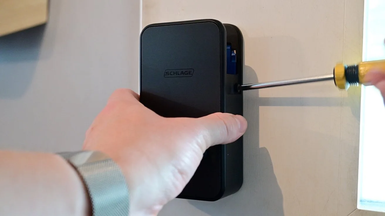 Person using a small screwdriver to tighten or loosen a screw on a rectangular black electronic device mounted on a wall, with one hand holding the device steady
