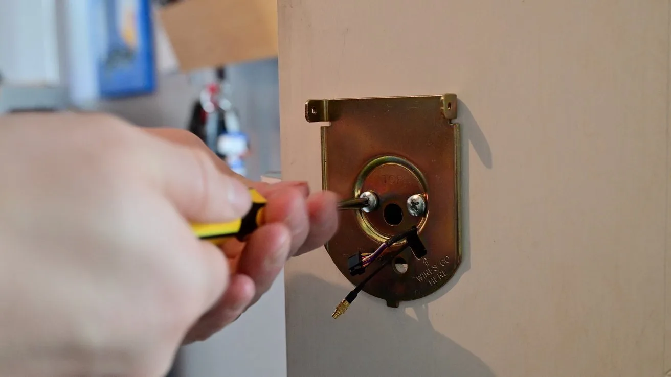 Hand using a screwdriver to install or remove an electrical wall fixture, with exposed wires and metal mounting plate on an indoor wall.