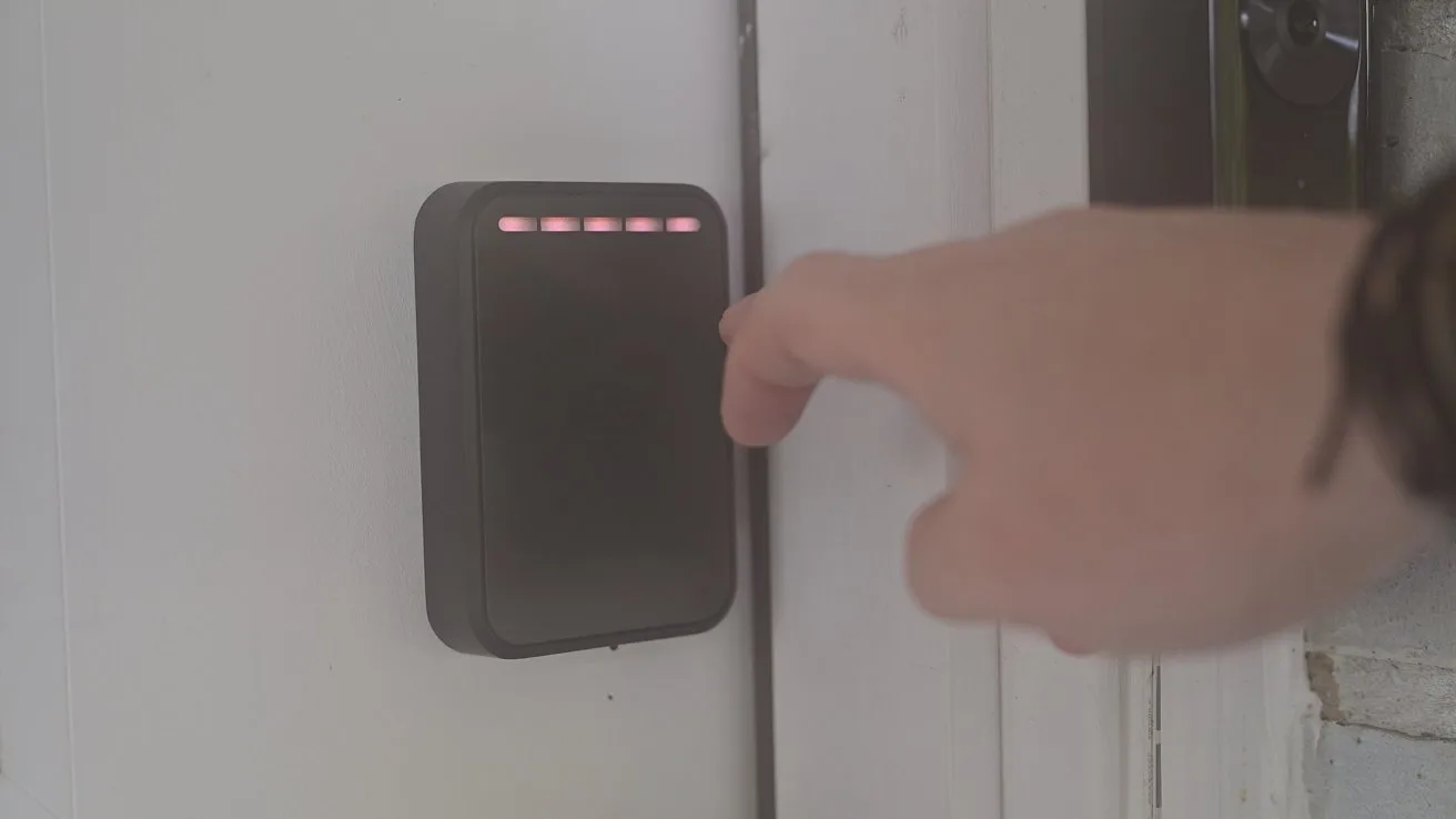 Hand reaching toward a wall-mounted electronic access panel with small red indicator lights, next to a closed door indoors