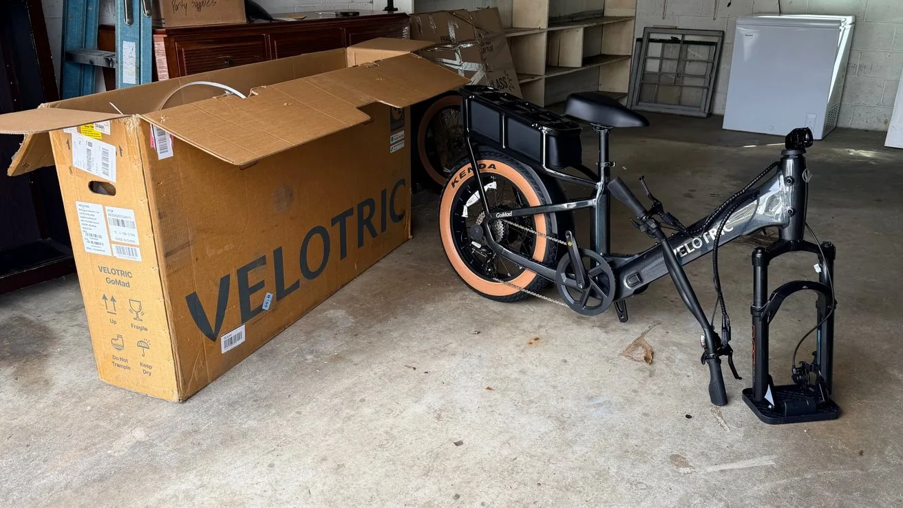 Partially assembled black Velotric electric bicycle with tan tires on a garage floor beside a large open Velotric shipping box and detached front wheel with fork and fender
