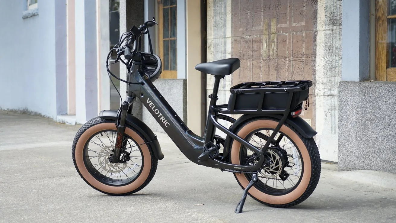 Black Velotric electric bike with tan fat tires, step-through frame, rear cargo rack, and kickstand, parked on a sidewalk in front of a building with windows and textured walls