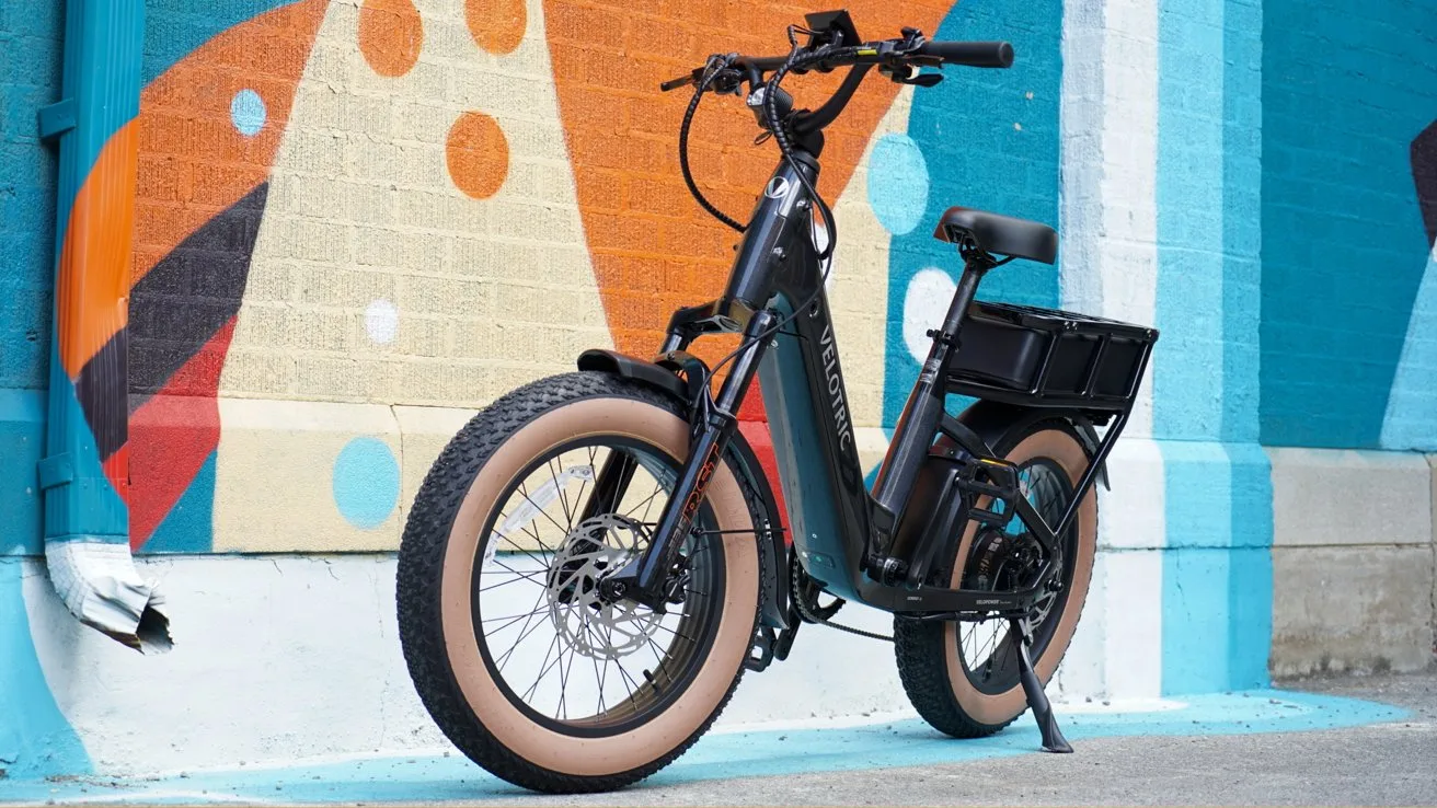 Black Velotric electric cargo bike with fat tan tires parked on a sidewalk in front of a colorful abstract mural featuring orange, blue, and white geometric shapes and polka dots