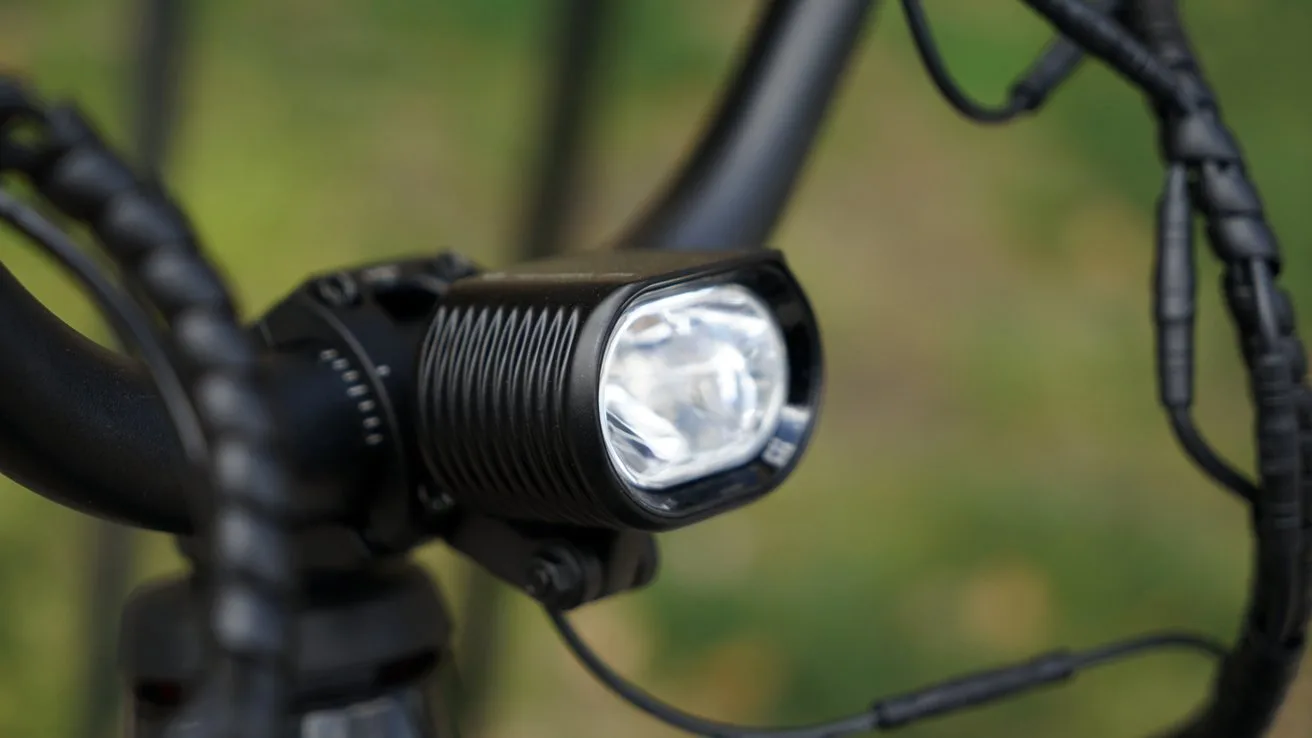 Close-up of a black bicycle handlebar-mounted headlight with ridged housing and bright lens, surrounded by coiled cables, against a blurred green outdoor background