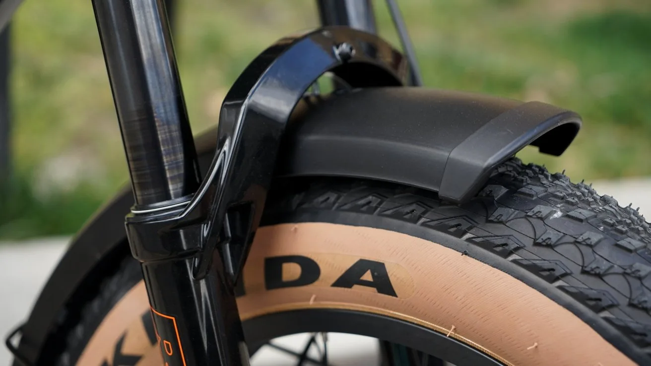 Close-up of a bicycle front wheel with tan sidewall knobby tire, black suspension fork, and curved black plastic fender, outdoors with blurred green grass background