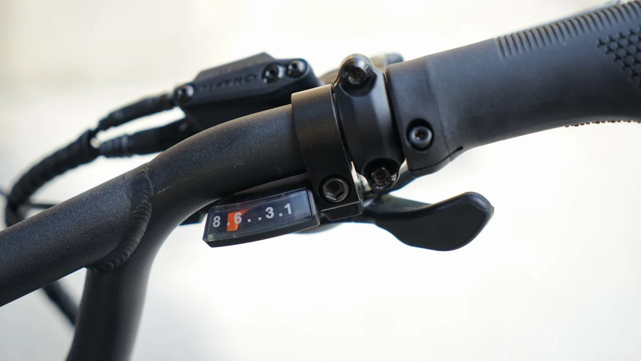 Close-up of a black bicycle handlebar showing textured rubber grip, brake lever, gear shifter, and a small gear indicator window displaying numbers 8, 6, 3, and 1 against a light background
