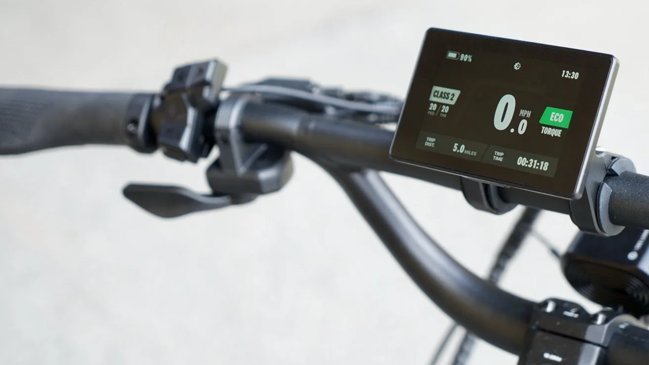 Close-up of an electric bicycle handlebar with ergonomic grips, control buttons, and a digital display showing speed, battery level, eco mode, trip distance, and ride time against a blurred background