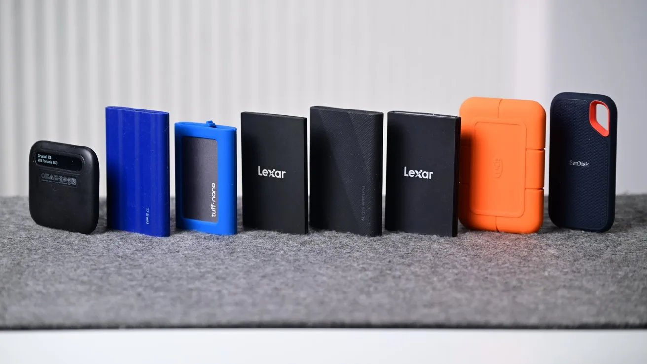 Row of seven portable external hard drives and SSDs in various colors and shapes standing upright on a gray surface against a softly blurred light background