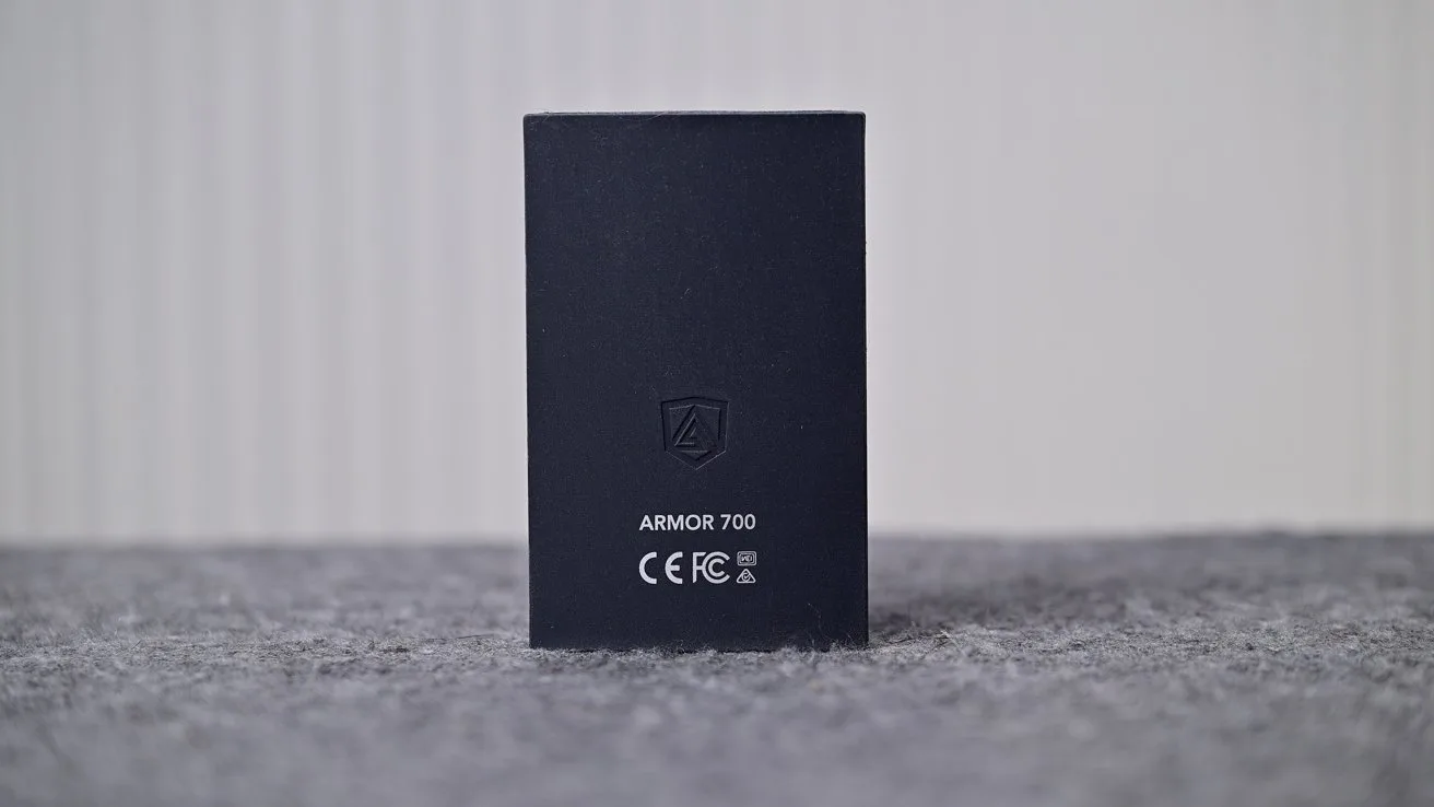 Black rectangular electronic device standing upright on a gray textured surface, showing a shield logo, the text ARMOR 700, and certification symbols against a softly blurred light background