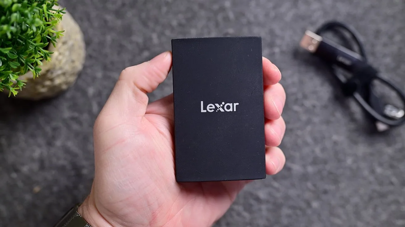 Hand holding a small black Lexar external drive over a gray surface, with a coiled USB cable and a small green potted plant in the background