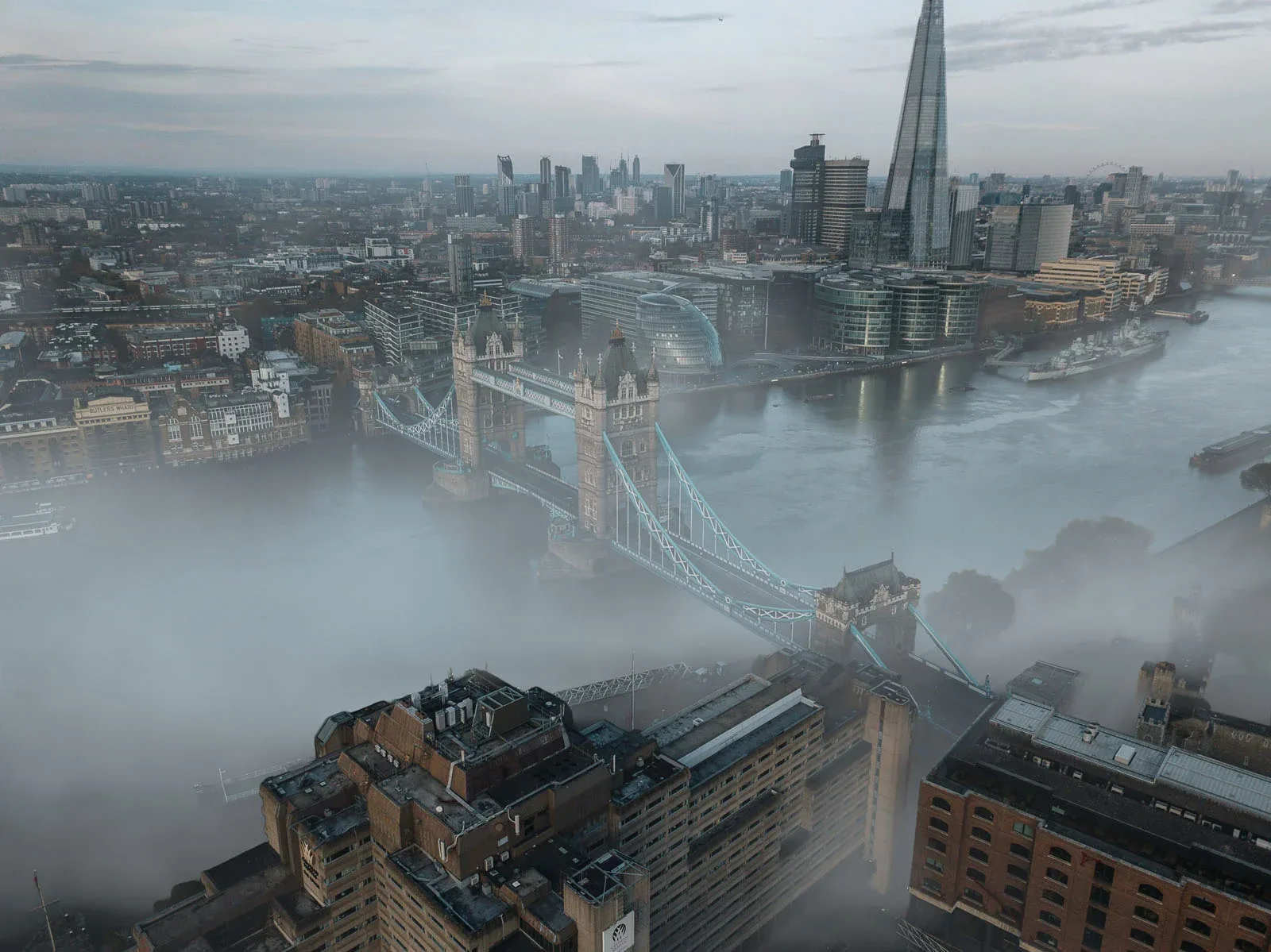 An aerial view of London's Tower Bridge spanning the River Thames, partially obscured by thick morning fog.