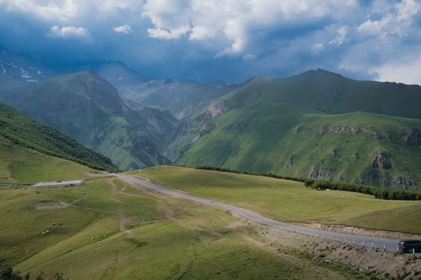 A paved road winds through a vast, green mountain valley under a dramatic, cloudy sky.